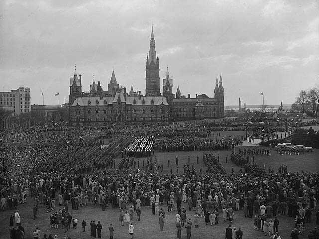 Black and white photograph. Thousands of people gather near the parliament buildings in Ottawa. A stage is set up in the middle right of the photo, several vehicles are nearby.
